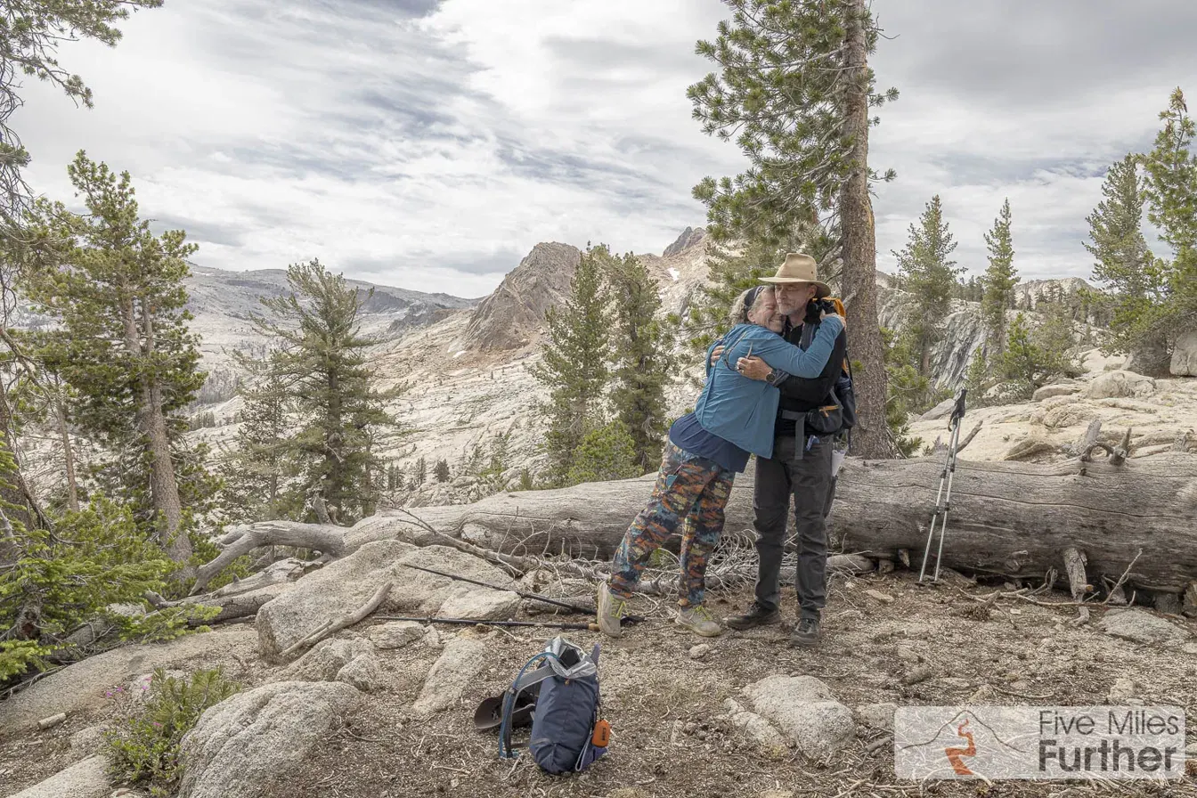 Two hikers embracing at a high mountain overlook with peaks behind them